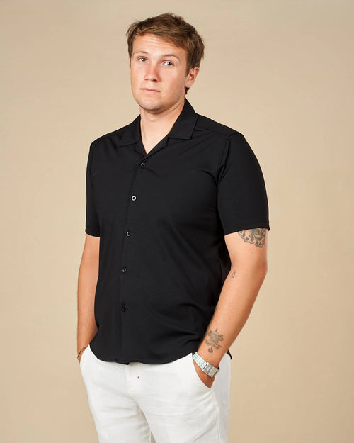 Man wearing a black Cuban collar shirt untucked with white trousers