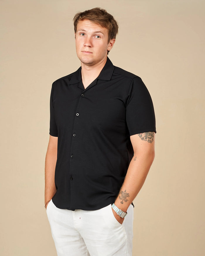 Man wearing a black Cuban collar shirt untucked with white trousers