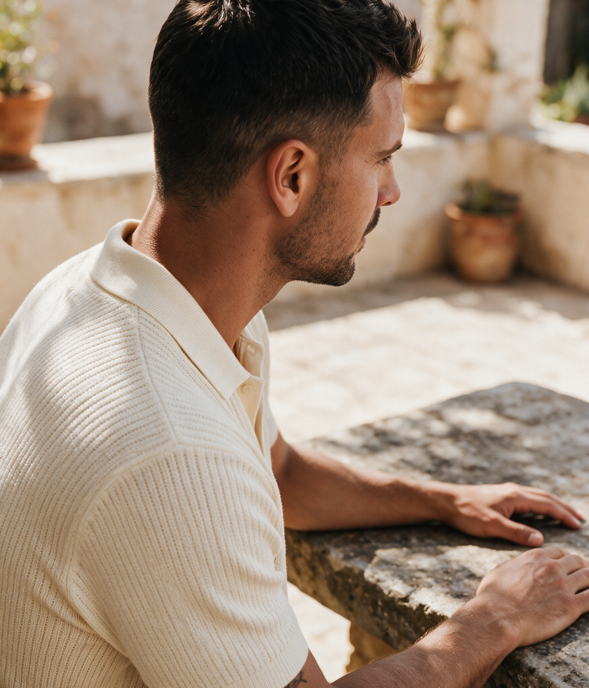 Man wearing a cream ribbed knitted polo shirt seated in a sunlit Mediterranean courtyard — Legacy London