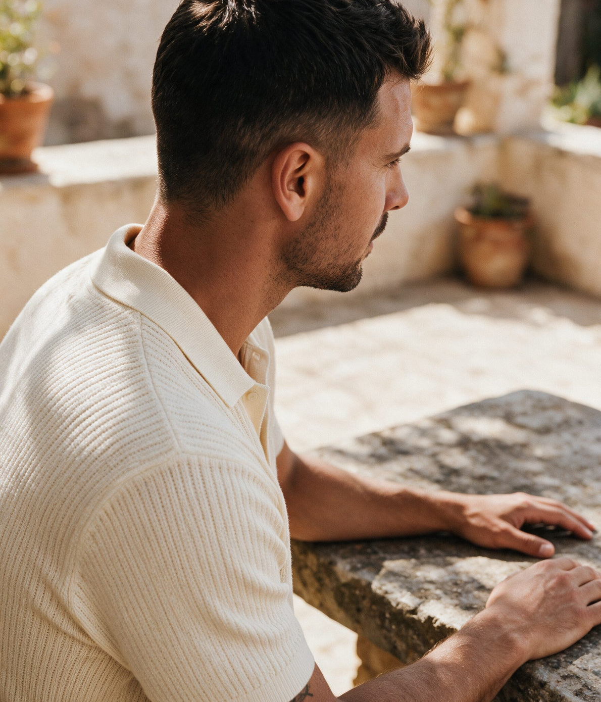 Man wearing a cream ribbed knitted polo shirt seated in a sunlit Mediterranean courtyard — Legacy London